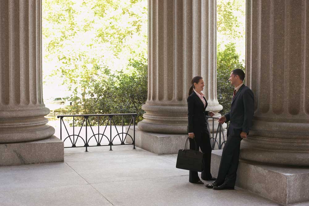 Image of two lawyers in an outdoor rotunda of a courthouse, smiling collegiately to one another during a discussion depicting the concept of attorney camaraderie.