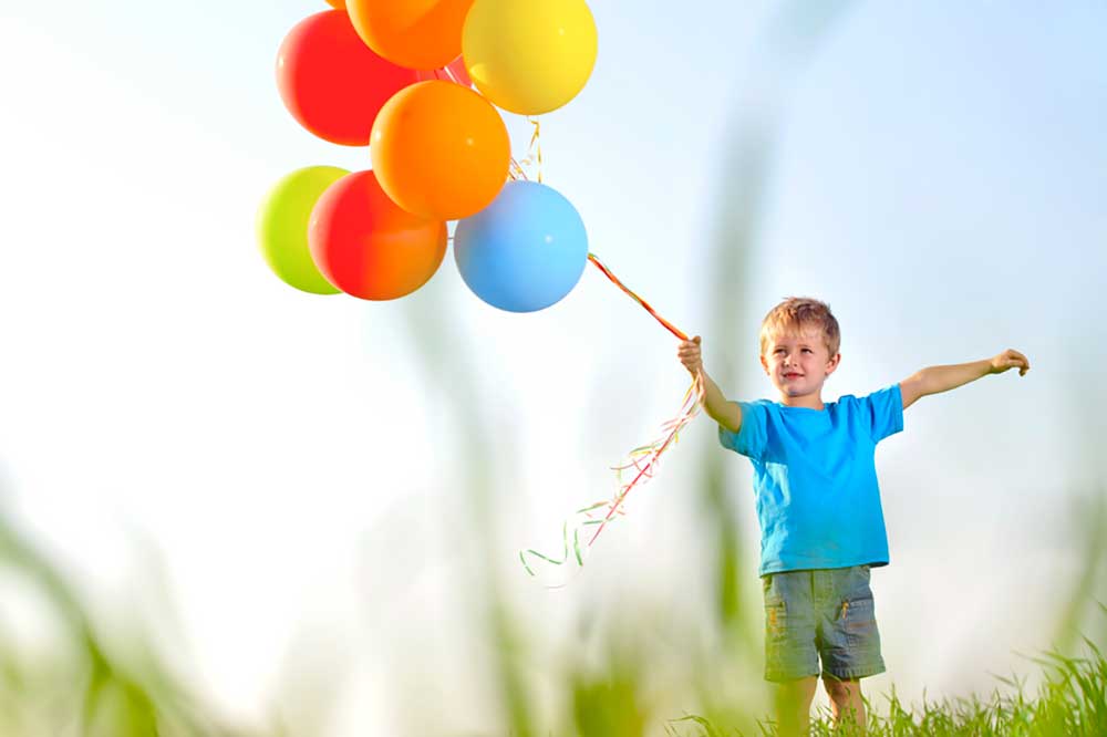 Picture of a small boy in a field of grass holding a cluster of brightly colored helium balloons on a string
