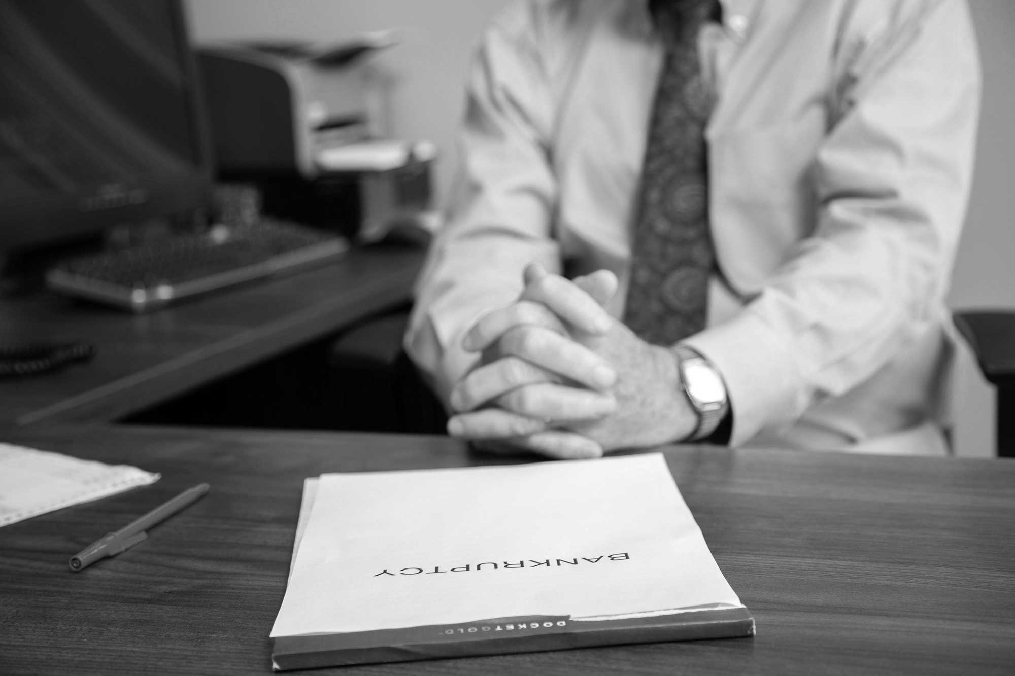 Bankruptcy Image of Mr. Hillier behind his desk with his hands folded. On his desk in front of him is a folder labeled bankruptcy.