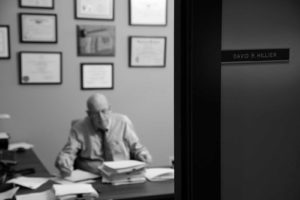 Photo close up of David R. Hillier's name plaque on the outside of his office door. Mr. Hillier himself sits in the back ground at his desk, working.