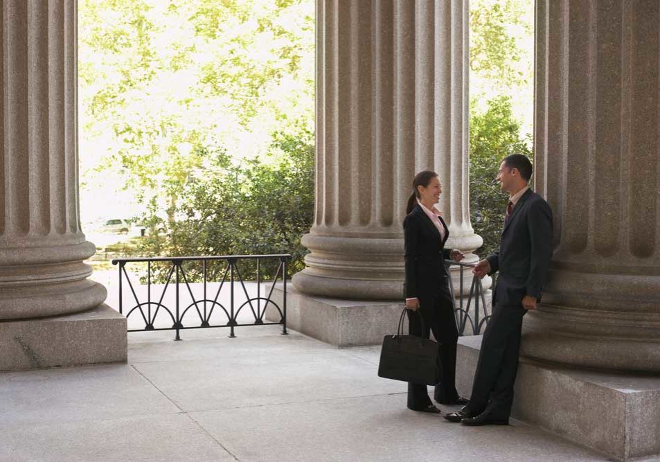 Lawyers after the Hearing Image of two lawyers in an outdoor rotunda of a courthouse, smiling collegiately to one another during a discussion depicting the concept of attorney camaraderie.