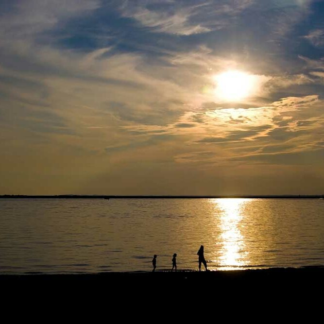 Single parent and two small children walking on the distant beach at dusk in silhouette.