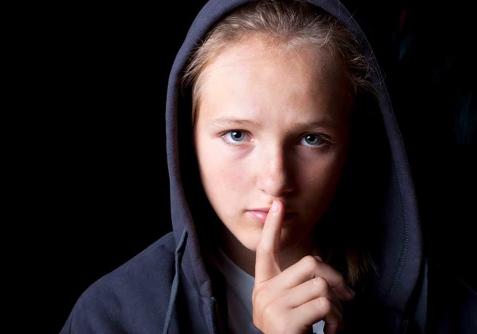 Domestic Violence Image of a child wearing a black hoodie on a black background with his finger in front of his lips illustrating saying nothing.