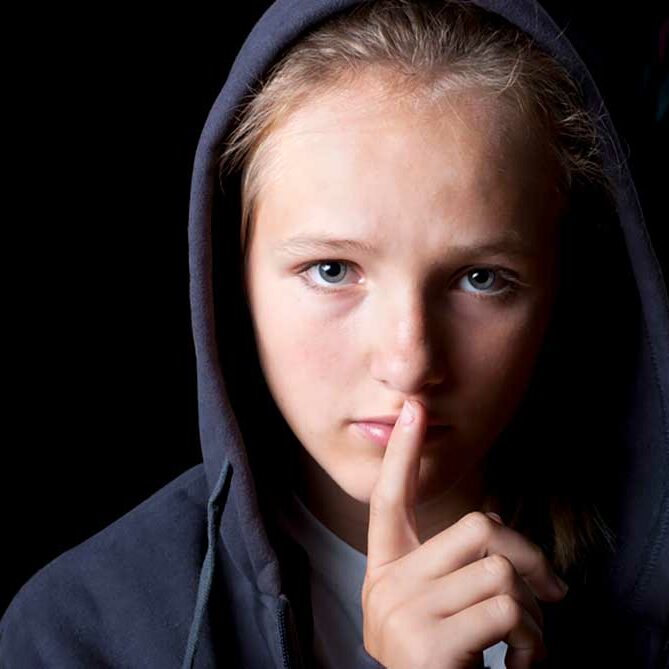 Image of a child wearing a black hoodie on a black background with his finger in front of his lips illustrating saying nothing.