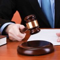 Image of a man in a black suit and blue tie, cropped without a face, pounding a gavel.