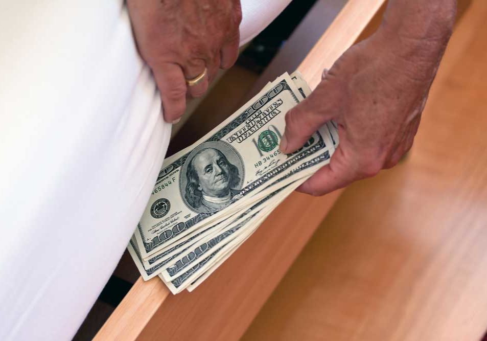 Hidden and Missing Assets Picture of a man's hands wearing a wedding ring hiding money under the mattress.