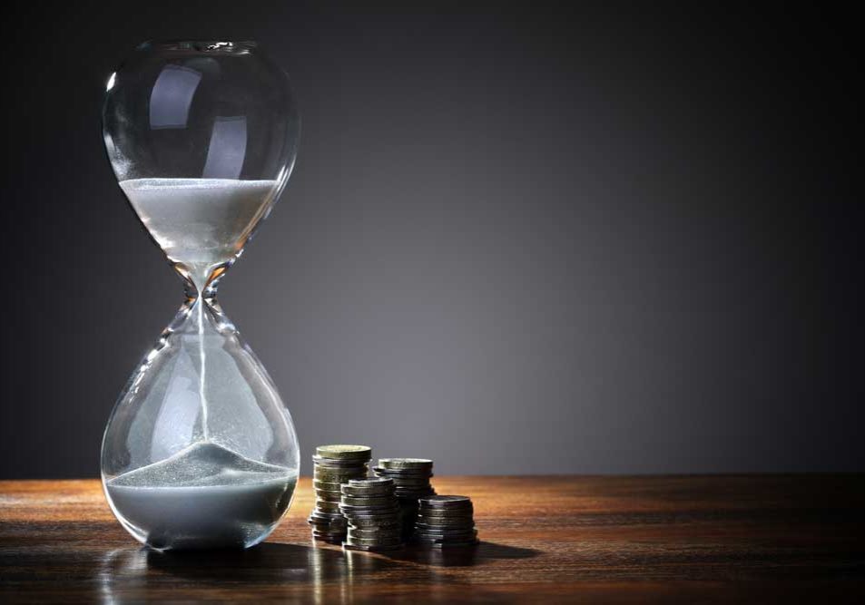 Time & Money Image of an elegant hourglass on a wooden table in front of a gray wall, representing time and money.