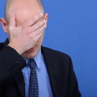 Image of man standing on a blue background with his hand over his forehead as if ashamed of the mistake he has made.