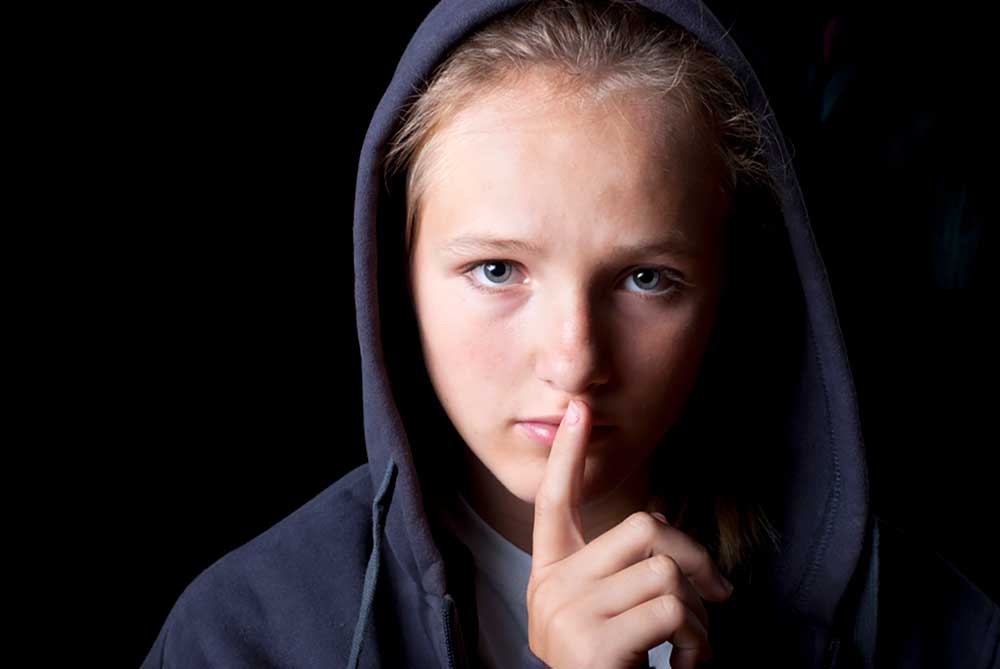 Image of a child wearing a black hoodie on a black background with his finger in front of his lips illustrating saying nothing.