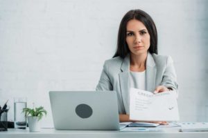 Image of woman sitting behind a desk with a laptop between her and the foreground, holding out a credit report document to an unseen recipient.