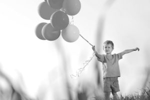 Black and white stock photograph of a small boy holding wind-blown balloons on a field denoting a healthy, happy child.