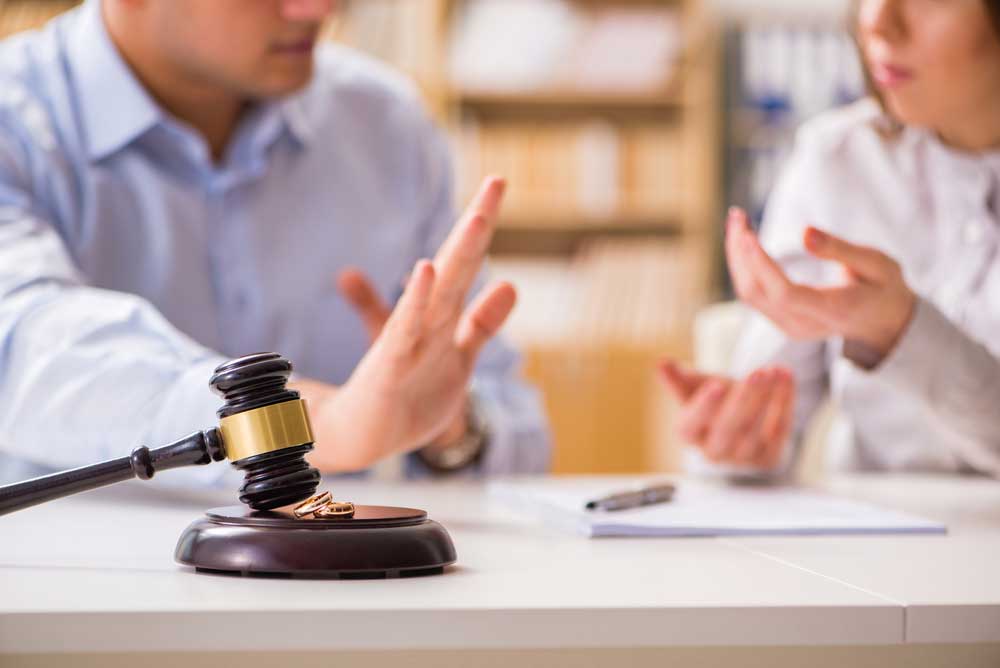 Image of two people sitting at a desk cropped so that faces are implied. The gavel in the foreground denotes a legal proceeding and the hand gestures of the subjects implies that one spouse disagrees.