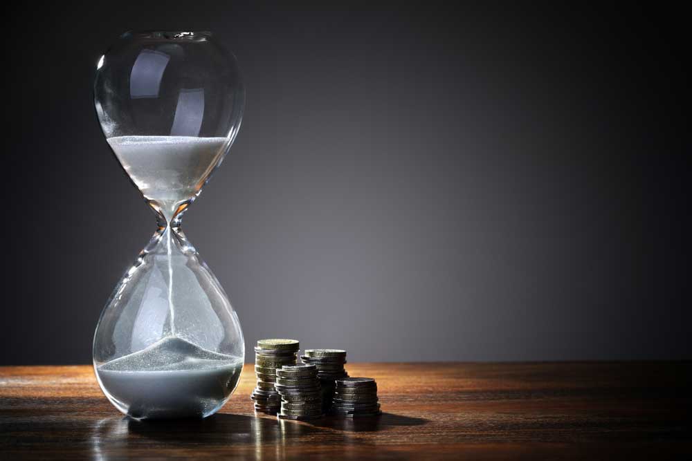 Image of an elegant hourglass on a wooden table in front of a gray wall, representing time and money.