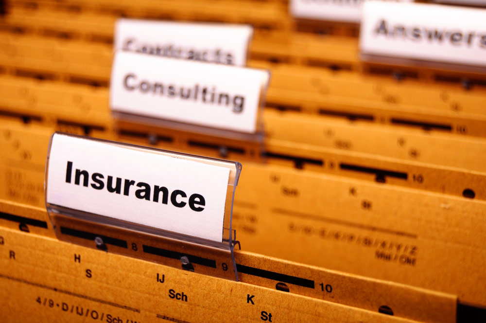 Image taken from across the top of a filing cabinet drawer with hanging file folders with plastic tab separators. The foremost forward tab is labeled "Insurance" while remaining tabs are slightly out of focus.