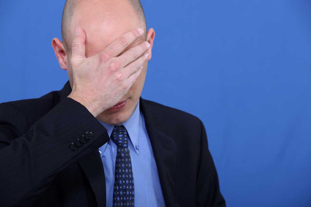 Image of man standing on a blue background with his hand over his forehead as if ashamed of the mistake he has made.