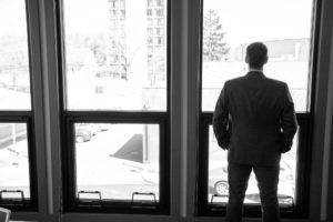 Black and white image of Patrick McCroskey, standing back to the camera, looking out his office windows.