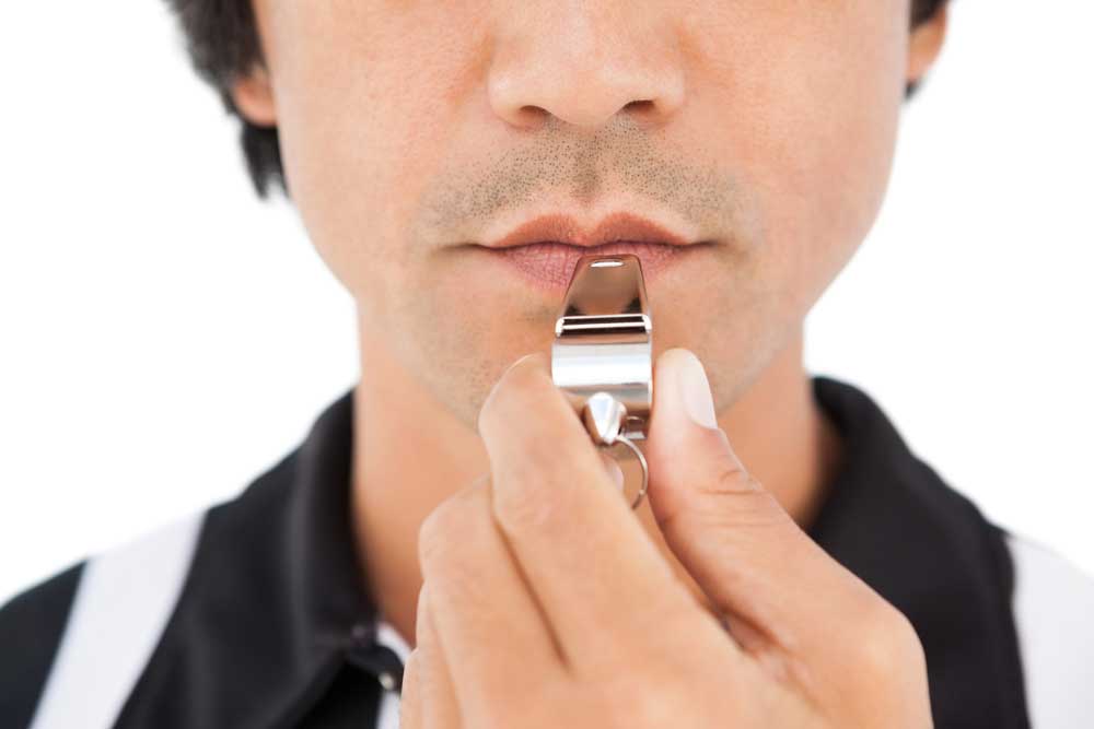 Cropped image of the bottom of a referree's face and top of his shoulders holding a whistle to his mouth