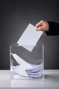 Image of a person dropping a card into a clear glass container, like a suggestion box or ballot bowl.