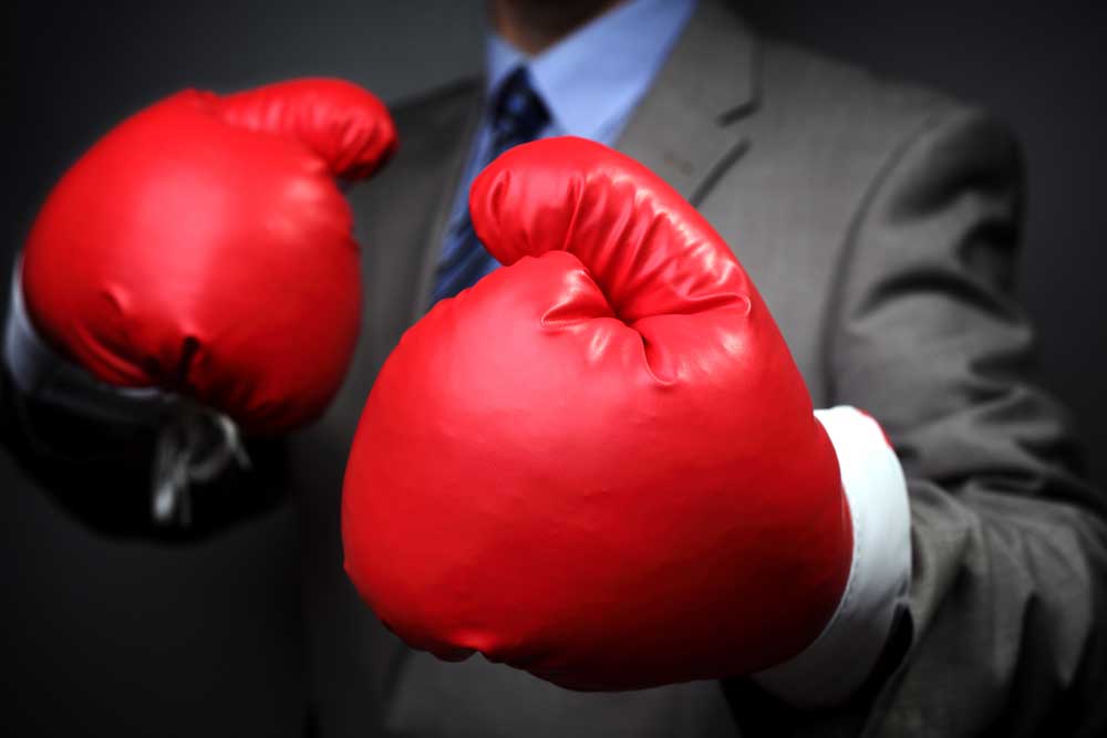 Image of a man in a gray suit on a dark background wearing red boxing gloves close to camera.